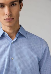 Camicia azzurro chiaro con bottoni, caratterizzata da un motivo testurizzato di piccoli punti scuri, colletto aperto e chiusura con bottoni standard. Vestibilità aderente.