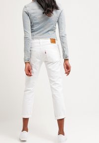 Donna in piedi con le spalle rivolte verso di noi, indossa una leggera giacca di denim, jeans bianchi arrotolati e sneakers bianche, su uno sfondo neutro.