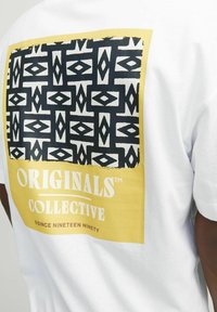 T-shirt bianco in cotone con una grande grafica sul retro. Il design include un motivo geometrico nero all'interno di un rettangolo giallo e testo bianco.