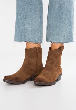 Brune ruskinds ankelstøvler med sidelukning, båret med lyseblå jeans med frynset kant på hvid baggrund.