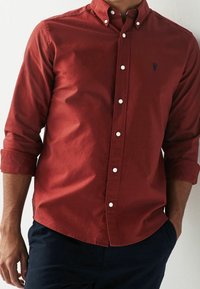 Camisa roja abotonada con mangas largas, confeccionada en un tejido texturizado y botones blancos, complementada con un pequeño logo azul marino en el pecho.