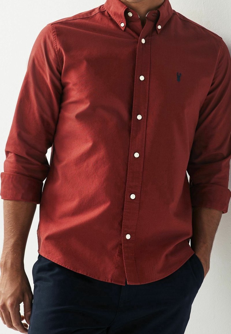 Camisa roja abotonada con mangas largas, confeccionada en un tejido texturizado y botones blancos, complementada con un pequeño logo azul marino en el pecho.