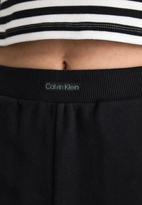 Juodos Calvin Klein kelnės su elastingu juosmeniu, su logotipo etikete. Viršuje matoma juodai baltos spalvos dryžuota trumpa palaidinė.