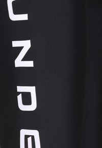 Czarne legginsy sportowe z wyraźnym białym napisem wzdłuż jednej strony. Tkanina jest gładka i elastyczna, zapewniając dopasowany kształt.