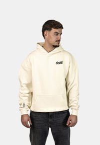 Hoodie color crema realizzato in tessuto morbido, con tasca frontale, cappuccio con coulisse e un piccolo logo nero sul petto e sulla manica.