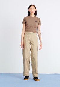 Camicia marrone a maniche corte con un piccolo logo, abbinata a pantaloni larghi color beige e sneakers nere con suole in gomma, caratterizzate da una texture liscia.