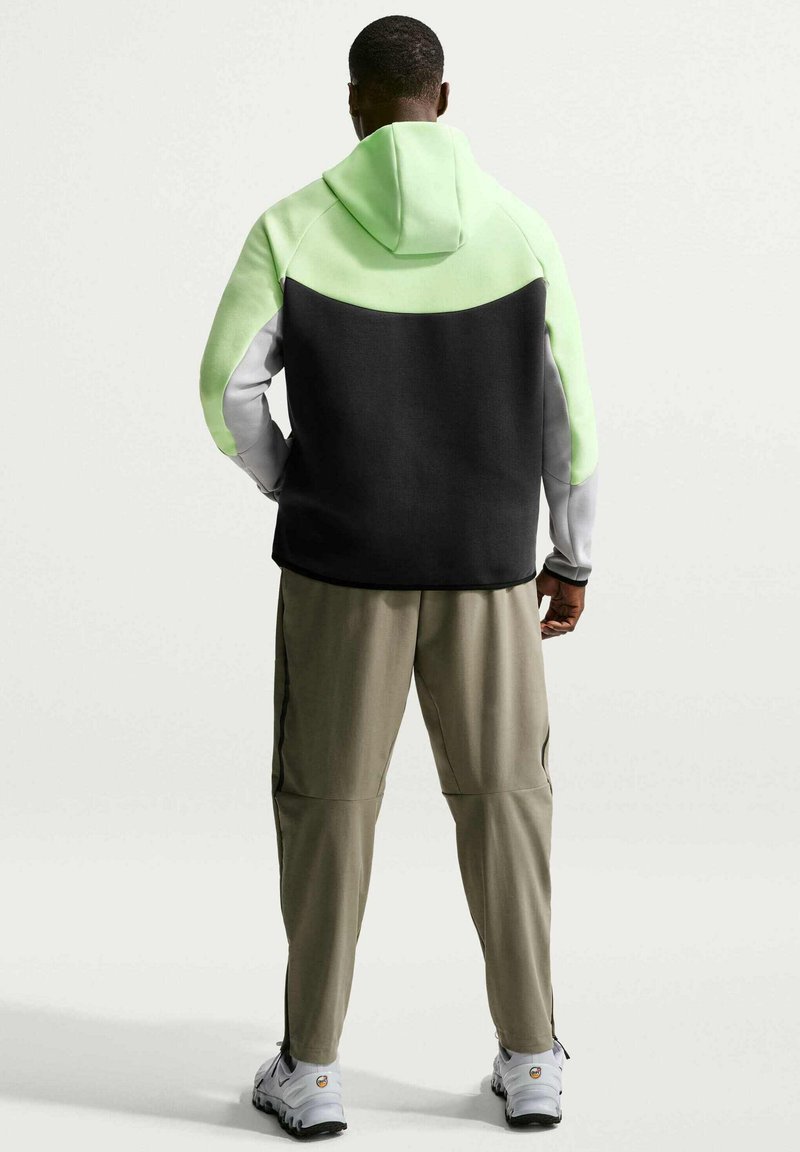 Mand stående med front mod væggen iført en color-block hættetrøje med limegrønne, sorte og grå paneler, beige bukser og hvide sneakers.