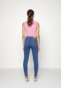 Jeans de mezclilla azules ajustados con cintura alta, bolsillos traseros y una etiqueta de cuero marrón; combinados con un top sin mangas rosa y tacones claros.