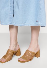 Brązowe plecione mule z otwartym czubkiem na słupku. Posiadają teksturowaną powierzchnię i kwadratowy kształt noska, w połączeniu z jasnoniebieską spódnicą dżinsową.