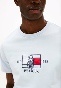 Camiseta azul claro de cuello redondo, con logo de Hilfiger bordado y un velero estilizado dentro de un diseño rectangular.