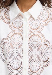 Blouse blanche à boutons, présentant des motifs de dentelle découpés intriqués, un col structuré et de délicats accents avec des textures à tissage ouvert.
