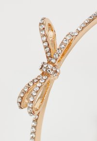 Accessorio in metallo dorato con design a fiocco, impreziosito da strass trasparenti lungo i bordi e al centro, che mette in evidenza i suoi intricati dettagli.