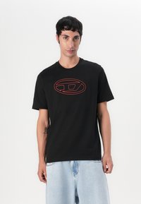 Svart bomulls t-skjorte med en rød oval logo-design på brystet. Korte ermer og rund hals. Modellen har på seg lyseblå denimshorts.