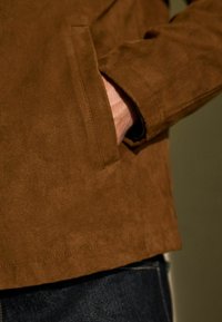 Chaqueta de ante marrón con una textura suave, que cuenta con un bolsillo lateral y un dobladillo recto; llevada sobre unos jeans de mezclilla oscuros.