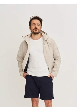 Man met baard, gekleed in een lichtbeige jas over een wit T-shirt en donkerblauwe korte broek, staand met handen in zakken tegen een effen achtergrond.
