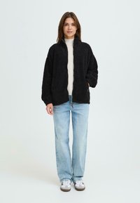 Svart, fuzzy jakke med høy krage, glidelås og sidelommer, kombinert med en lys genser og lyseblå jeans. Hvite joggesko.