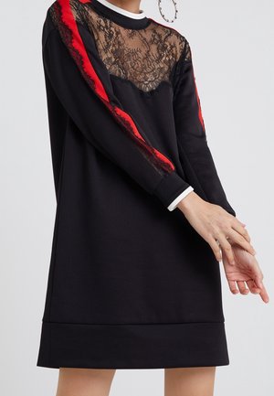 Robe noire avec un appliqué en dentelle sur la poitrine et des accents rouges le long des manches. Manches longues, col rond et coupe décontractée.