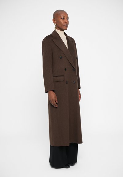Manteau en laine marron, à double boutonnage, avec des revers en pointe, des poches latérales et deux boutons noirs ; associé à un col roulé crème et un pantalon noir.