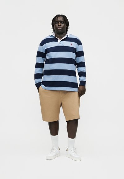 Egy férfi áll, világoskék és tengerészkék csíkos hosszú ujjú inget, bézs rövidnadrágot, fehér zoknit és fehér tornacipőt viselve, egyszínű háttér előtt.