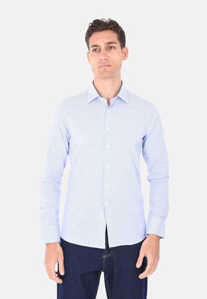 Uomo con camicia azzurra a righe con bottoni e jeans blu scuro, in piedi davanti a uno sfondo chiaro e uniforme.