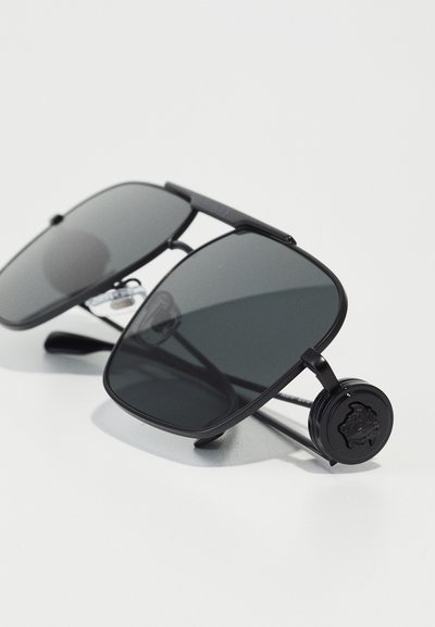 Lunettes de soleil noires avec des verres sombres et une forme rectangulaire. Accents en métal prononcés et un emblème circulaire de marque sur le côté.