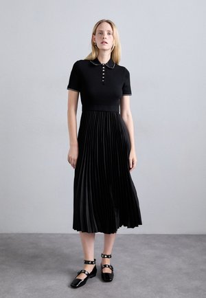 Vestido negro con un top acanalado, mangas cortas y una falda plisada. Presenta un cuello y detalles de botones. Acompañado de zapatos planos negros.