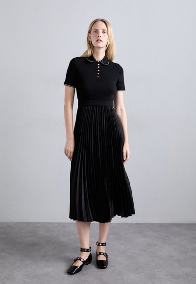 Robe noire avec un haut côtelé, des manches courtes et une jupe plissée. Dotée d'un col et de détails de boutons. Accompagnée de chaussures plates noires.