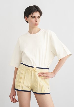 Kvinde med kort mørkt hår iført en oversized hvid T-shirt og blege gule strikkede shorts med marineblå kant og snøre.