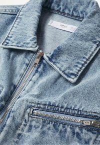 Veste en jean bleu clair avec une fermeture éclair argentée à l'avant, un col et une poche poitrine horizontale zippée.