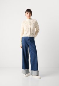 Cremfarbener, strukturierter Strickcardigan mit tailliertem Schnitt, kombiniert mit hochgestellten, weit geschnittenen blauen Jeans aus Denim mit ausgefransten unteren Säumen.