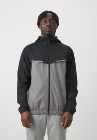 Veste imperméable en noir et gris. Dotée d'une fermeture éclair à l'avant, de poignets élastiques et d'accents réfléchissants. Légère avec un design épuré.