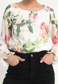 Blouse fleurie à base blanche, avec des motifs floraux roses et verts, des poignets côtelés et un col rond ; associée à un jean noir.