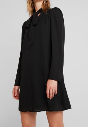 Woman wearing a loose black long-sleeve dress with a front bow tie neckline and hoop earrings against a plain light background.