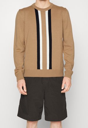 Man met een beige gebreide trui met verticale zwarte en witte strepen in het midden en zwarte knielange korte broek.