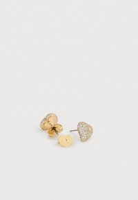 Zlaté náušnice v tvare srdca zdobené priehľadnými kryštálmi, s textúrovaným zadkom a malou visačkou s vyrytým logom značky.