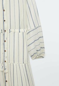 Robe rayée en crème et bleu, fabriquée en tissu léger. Présente des boutons sur le devant, des poignets élastiqués et un ourlet en cascade.