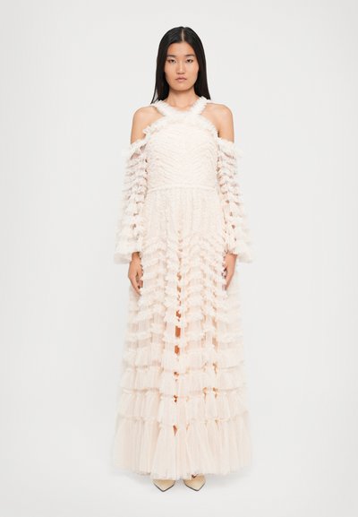 Woman in a long, off-shoulder, tiered cream dress with ruffled textured fabric, standing straight against a plain white background.