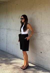 Mustad kõrge vöökohaga mustad šortsid, millel on sidumisvöö, koos sobiva valge kampsuniga ja mustade prillidega. Tekstuuriga seina taust.