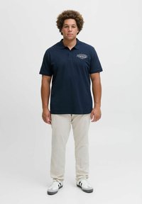 Marineblaues Poloshirt mit Kragen und kurzen Ärmeln, mit einem bedruckten Logo. Kombiniert mit hellfarbenen Hosen und weißen Sneakern.