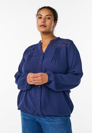 Ženska z kodrastimi lasmi, oblečena v mornarsko modro bluzo z čipkastim detajlom na ramenih, stoji z rokami zloženimi in pogumno gleda naprej.