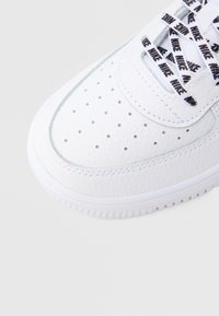 Biela kožená teniska s perforovaným dizajnom, textúrovaným povrchom, bielou podrážkou a čiernymi šnúrkami s logom. Obsahuje prešívané detaily.