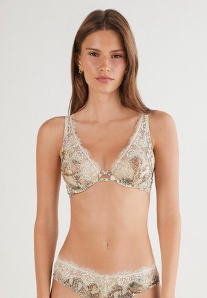 Vrouw in een beige kant en dierenprint beha en bijpassende slip met kant, staand tegen een effen lichte achtergrond.