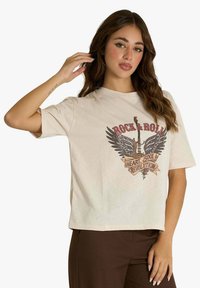 T-shirt beige con una grafica raffigurante una chitarra, ali e testo. Maniche corte, vestibilità comoda e scollo rotondo.