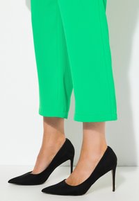 Černé nubukové lodičky s špičatou špičkou, spárované s živě zelenými zkrácenými kalhotami, které ukazují hladkou texturu a elegantní design.