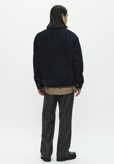 Námořnická kordurová bunda s volným střihem a knoflíkovými manžetami, doplněná o vrstvenou béžovou košili. Tmavě šedé kalhoty a černé boty.