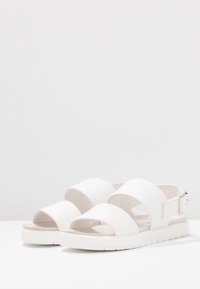 Bílé sandály se třemi širokými pásky, strukturovaným svrškem, otevřenou špičkou a nastavitelnou přezkou na patě. Silná bílá gumová podrážka.