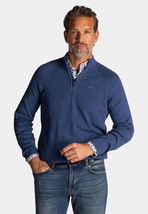 Man met baard draagt een blauwe sweater met rits tot aan de borst over een overhemd met patroon en blauwe spijkerbroek, staand met één hand in de zak, neutrale achtergrond.