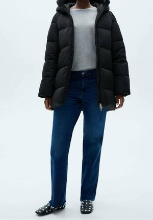 Personne portant une veste matelassée noire, un pull gris clair, un jean bleu avec des fentes sur les côtés, et des mocassins noirs ornés de clous argentés, se tenant devant un fond uni.