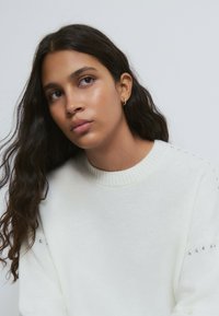 Junge Frau mit langen dunklen Haaren, die einen weißen Pullover mit kleinen silbernen Nieten trägt und leicht nach oben und links blickt vor einem schlichten Hintergrund.