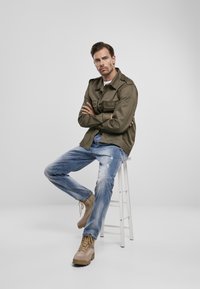 Chaqueta verde oliva de estilo militar, camiseta blanca, vaqueros azul desgastados, botas marrón claro. Modelo sentado en un taburete blanco con los brazos cruzados.